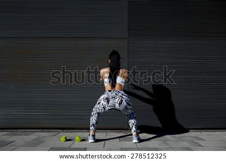 Back view athletic woman with perfect figure and buttocks doing squats against wall with copy space for your text message,fit female in sportswear squatting after training on black background outdoors