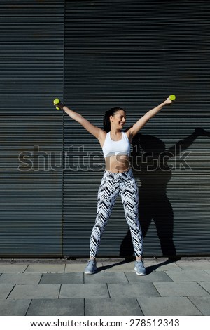 Smiling female with perfect figure getting her arms in great shape while lifting weights, attractive young woman using dumbbells to work out her arms while training outdoors on copy space background