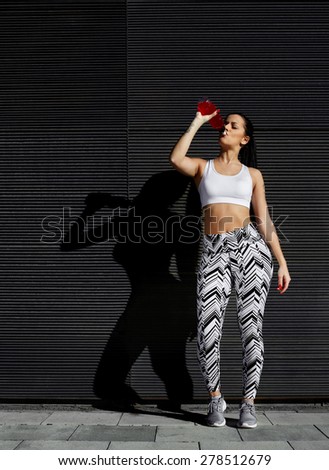 Young fit woman with beautiful figure refreshing with energy drink against black wall in the city,female runner taking break after workout standing against background with her amazing feminine shadow