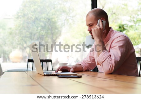 Handsome business man talking on smart phone and look to laptop screen, male freelancer working on computer at wooden table of modern coffee shop loft, young hipster having conversation on cell phone