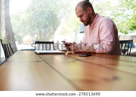 Male student sitting at wooden table of modern university library, handsome man using smart phone sitting in modern coffee shop with big windows, young business man text messaging at office desk