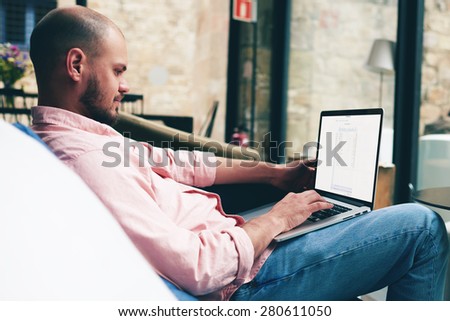 Relaxed young man sitting on sofa front open laptop computer while looking to the digital screen, successful freelancer using notebook in modern interior looking pleased and satisfied with his work