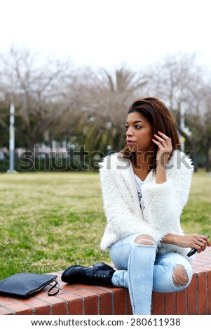 Portrait of trendy female hipster dressed in stylish clothing ready for tryst waiting for someone hold smart phone in the hand, young afro american woman sitting in beautiful park looking away
