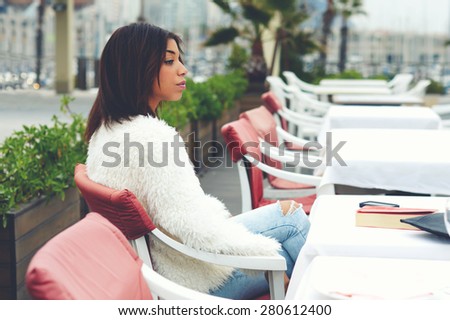 Portrait of attractive afro american woman enjoying a good day in restaurant while sitting at the table, charming female hipster sitting on the terrace of sidewalk cafe with palm tree on background