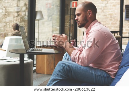 Male student working with laptop computer in modern university hall or library, young business man looking to the screen of open notebook while holding cup of coffee in modern loft interior, filter