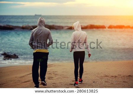 Two runners walking on the beach while they rest after intense morning training, sporty couple of friends taking break after workout outdoors at seashore with beautiful orange sunrise on background