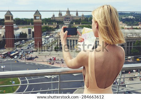 Young woman in dress photographing urban view with mobile phone camera during summer journey, female tourist taking picture of active life in the city with her smart phone while standing on viewpoint