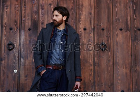Handsome confident mature man with beard stands on wooden brown background with copy space for your text message or advertising, fashionable rich male dressed in expensive clothes posing outdoors