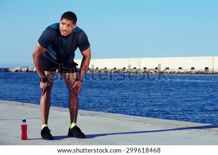 Tired male runner took a break after hard workout outdoors standing on sea and sky background with copy space area for your text message or information, sweaty sportsman having rest leaning on knees