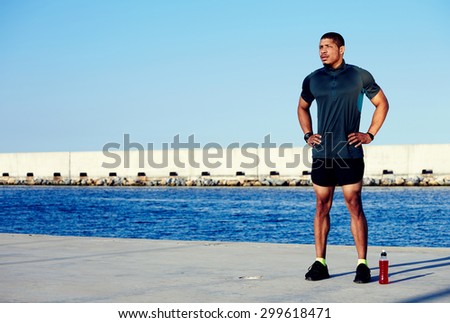 Strong pumped body man resting after workout in marina port while standing against the sea and sky background with copy space area for your text message or information, male jogger rest after running