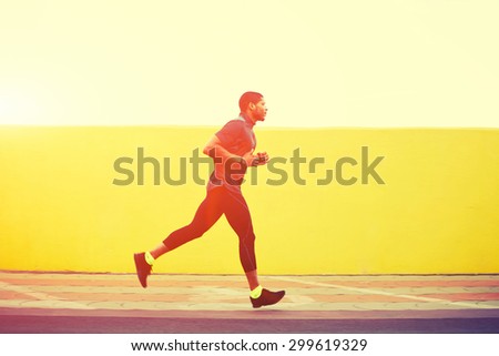 Full length portrait of afro american runner with muscular body in tracksuit jogging against yellow wall with copy space area for your text message or content, young male jogger working out outdoors
