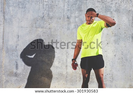 Tired male runner rubbing his forehead while resting after workout against cement wall background with copy space area for your text message or advertising, muscular athlete resting after fit training
