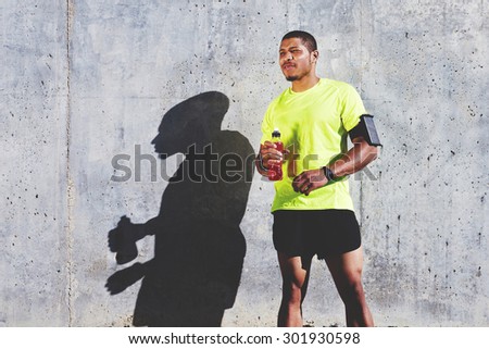 Afro american runner with muscular body in tracksuit relaxing with a bottle of energy drink while standing against cement wall with copy space area for your text message or content, athlete resting