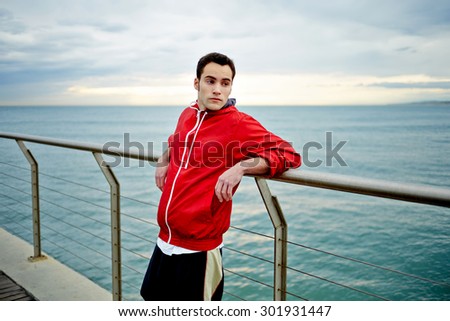 Portrait of male runner taking break after run while standing on a pier enjoying beautiful sea landscape view, sporty young man resting after active physical training outdoors, jogger taking break