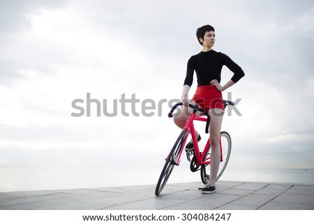 Portrait of stylish female hipster standing with her sport fixed gear bicycle against of cloudy dramatic sky background at seashore, cyclist and copy space area for text message or advertising content