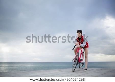 Young stylish female cyclist leaning on her light weight fixed gear bicycle while standing against copy space dramatic sky on seashore, sporty woman actively enjoying weekend strolling on her bike