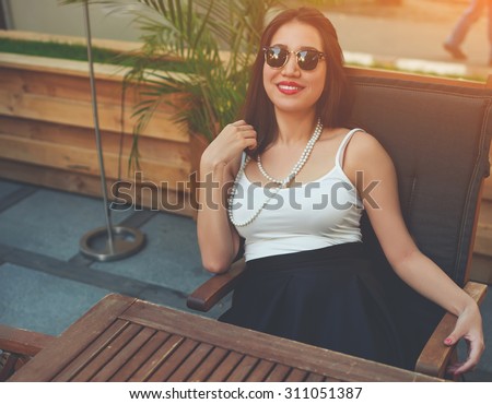 Portrait of a young beautiful woman in classy modern sunglasses with a bright smile on the face sitting in a cozy restaurant exterior terrace, fashionable female posing for the camera in sidewalk cafe