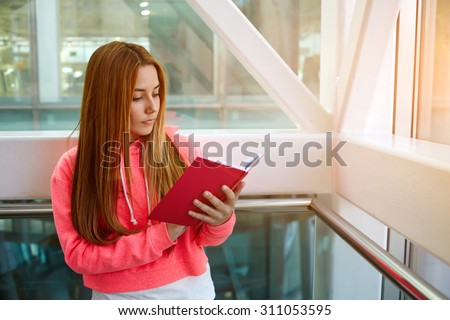 Attractive intelligent college student reading workbook or notebook while standing in the hallway of a modern building, stylish young hipster girl preparing for the exam at the university indoors