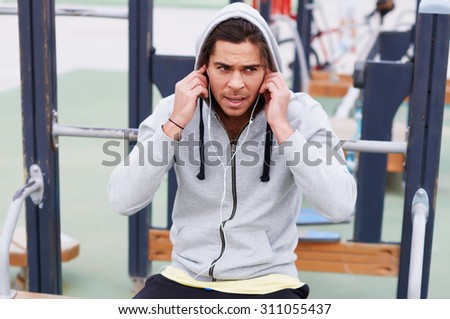 Exhausted handsome sportsman taking break after physical exercise outdoors while listening to music with headphones,tired young runner enjoying rest after hard workout outdoors on the horizontal bar