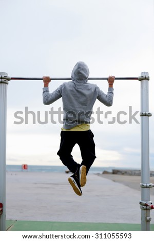Back view of a male runner doing pull ups on the horizontal bar while training at evening outdoors, strong athlete in tracksuit doing exercise at street gym apparatus, young sportsmen playing sports