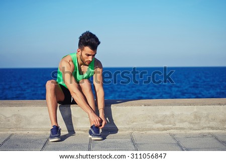Full length portrait young fit man sat on concrete pier to tie the laces while rest after run along beach, male runner taking break after fitness training outdoors with copy space area for your text