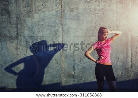 Charming woman holding hand at forehead while standing against of gray wall with its own shadow
