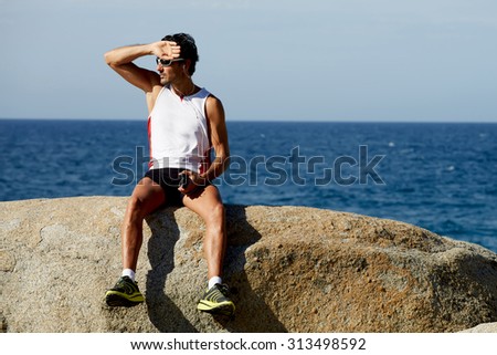Handsome male jogger listening to music in headphones and enjoying seashore landscape in sunny day, mature sports man taking break after active training outdoors while holding smart phone in the hand