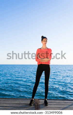 Full length portrait of female runner with a perfect figure having rest while standing on a wooden pier against blue sea and sky background with copy space area for text message or advertising content