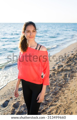 Portrait of a young attractive fit woman posing for the camera while standing on the beach in sunny summer day, female runner enjoying the rest after workout outdoors at beautiful seashore