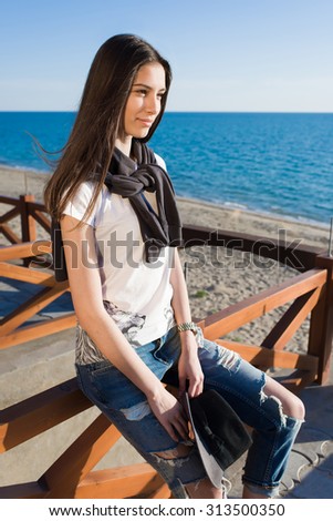 Charming young woman enjoying a good weather and recreation time while sitting against the sea in sunny afternoon, stylish female hipster rest after strolling along the beach in spring season