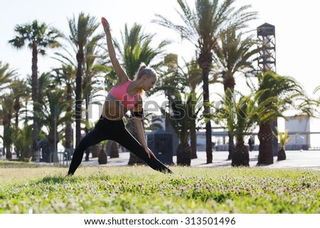 Full length portrait of fitness woman enjoying her outdoors warm up while listening to music in headphones, female runner stretching her body before begin morning run in the park in sunny summer day