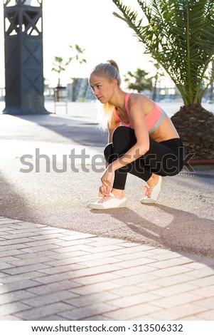 Beautiful female runner tying shoelaces on her jogging shoes while taking break after workout outdoors in a summer day, fit woman in sportswear resting after a morning jogging around the city