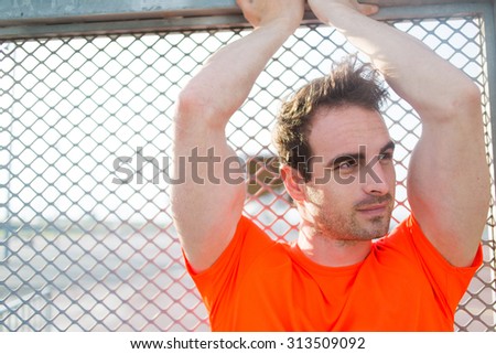 Portrait of attractive male jogger standing against urban metal fence while taking break after intense fitness training outdoors, caucasian muscular build man having a rest after workout at sunset