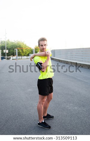 Young male runner stretching arms before start morning run in an urban setting, handsome athlete playing sports on the road at sunny morning while listening to music with headphones on his smart phone