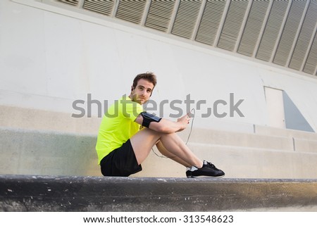 Attractive male runner in bright fluorescent t-shirt listen to music and having rest after morning training in urban setting, young sports man with mobile phone in the hand sits on steps taking break
