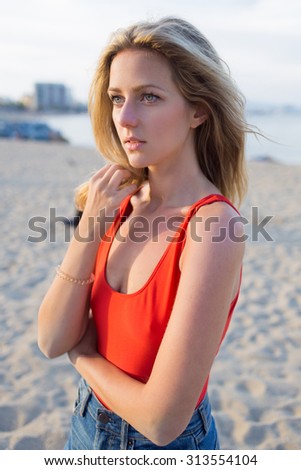 Portrait of young blonde hair charming woman standing alone on the beach waiting for someone, caucasian female with beautiful eyes posing for the camera near the sea in sunny summer evening