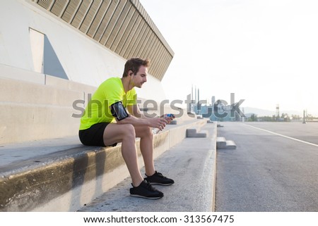 Young sports man holding bottle of water while taking break after an active run outdoors in early morning, male jogger listening to music in headphones while enjoying the rest after workout training