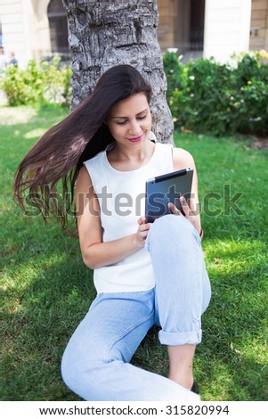 Young hipster girl reading fashion news on her touch pad while resting after strolling in summer day, attractive latin woman watching video on digital tablet computer while relaxing in the park
