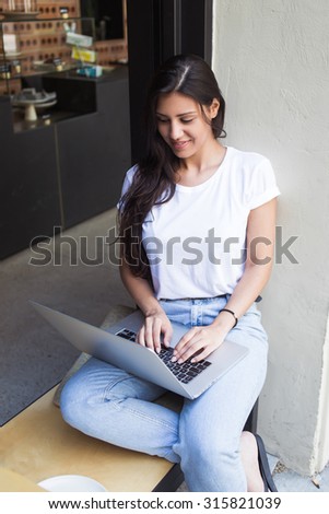Charming female student keyboarding on her net-book while resting in coffee shop after lectures, young latin woman using laptop computer for distance work while sitting in sidewalk cafe outdoors