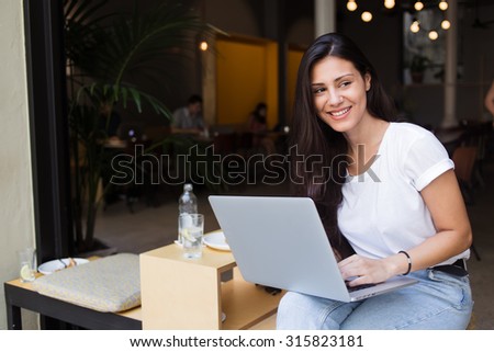 Portrait of a smiling hipster girl using laptop computer with copy space area for your text message or advertising content, charming latin women looking aside while holding on knees her open net-book