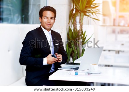 Successful freelancer man work remotely on his laptop computer while sitting at modern restaurant, confident businessman sitting at the table with open net-book and holding mobile phone in the hand