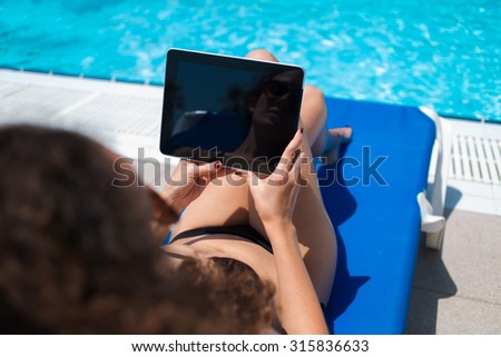 Cropped image of brunette woman reading e-book on touch pad while sunbathing near pool during weekend, female in bikini holding digital tablet with blank copy space screen for your advertising content