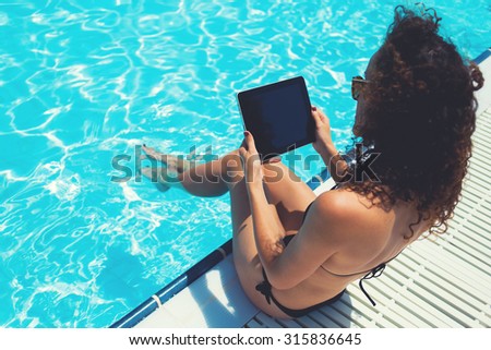Brunette hair woman in black bikini using touch pad for distance work during summer vacations, female holding digital tablet with blank copy space screen for your advertising while sunning near pool