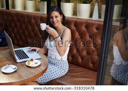 Satisfied successful businesswoman in elegant clothes sitting at cafe table with net-book enjoying a cup of cafe she smile and looks to the camera, young latin female at work break in a coffee shop