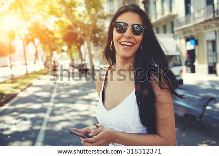 Happy Latin woman in summer sun glasses smiling brightly while holding smart phone strolling outdoors in the city at sunny day,female person using mobile standing with composition copy space on a side