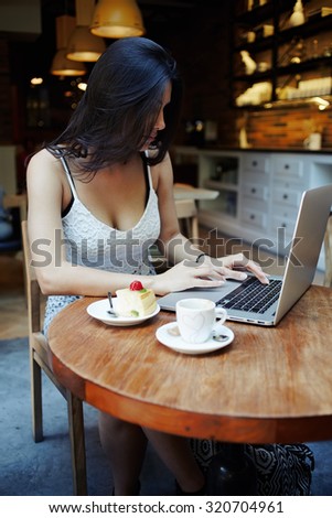 Portrait young elegant sexy woman with gorgeous breasts breakfast in luxurious coffee shop and work on laptop computer, female freelancer sit at the table with net-book, cup of coffee and tasty cake