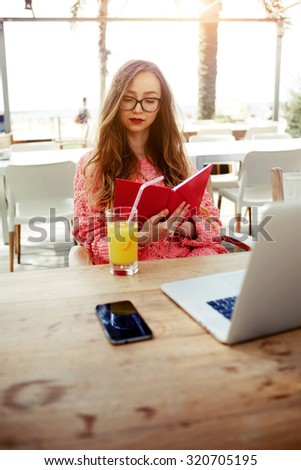 Attractive creative freelancer read notebook while sitting at wooden table with laptop laptop computer and cellphone, young female woman having breakfast on terrace in modern coffee shop, filter image