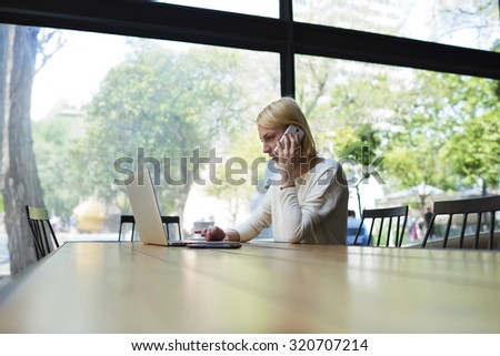 Female freelancer or student working busy on net-book, experienced young businesswoman keyboarding on her laptop computer while talking on smart phone sitting in modern office interior or coffee shop