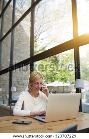 Female freelancer at distance work via internet talk on smart phone and looking to the net-book screen, young successful businesswoman busy working in modern loft office interior, flare sun light