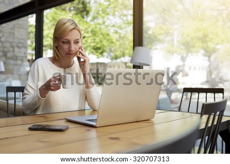 Female freelancer at distance work via internet enjoying her cup of coffee while talking on smart phone and looking to the net-book screen, young businesswoman at work break preparing for the meeting